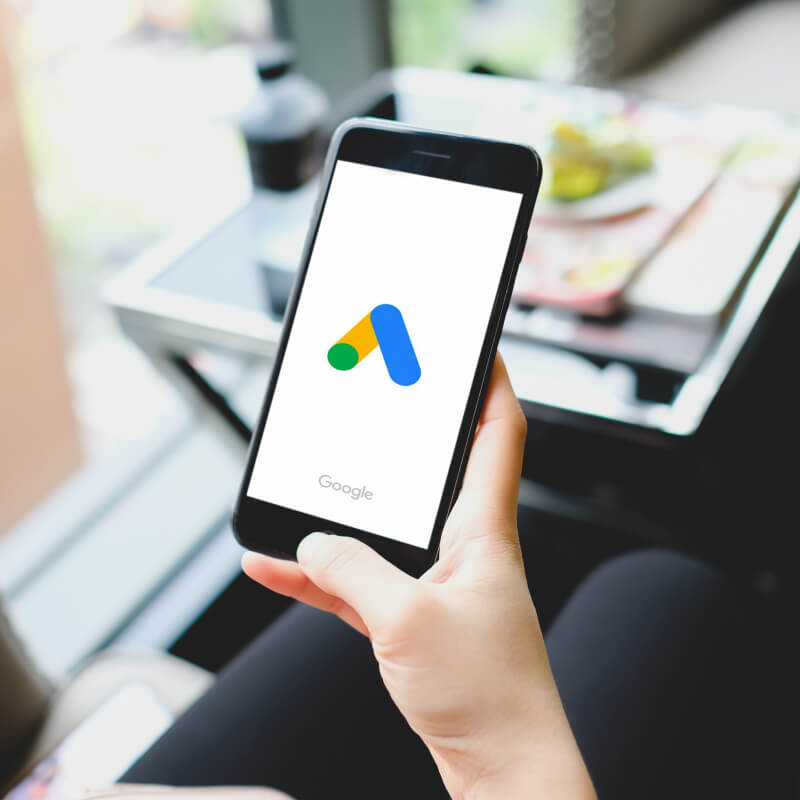 Telefon z logo Google Ads, trzymany w ręce, na tle stolika.