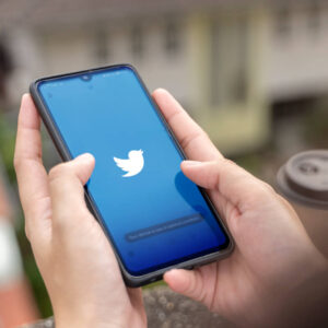 Osoba trzymająca w dłoniach telefon z logo aplikacji Twitter, z budynkiem w tle.