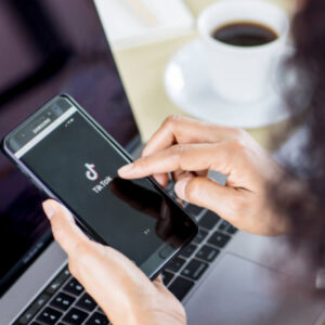 Osoba trzymająca w dłoniach telefon z logo TIkToka na ekranie, na tle biurka