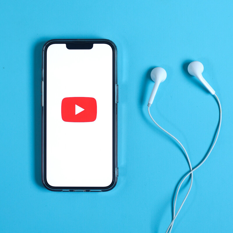 Telefon z logo YouTube na ekranie leżący obok białych słuchawek na niebieskim tle
