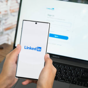 Telefon z logo LinkedIn na ekranie trzymany w dłoniach na tle laptopa