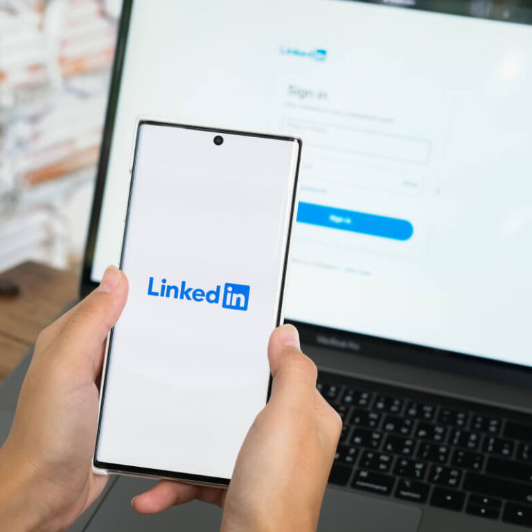 Telefon z logo LinkedIn na ekranie trzymany w dłoniach na tle laptopa