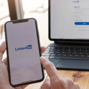 Osoba trzymająca w dłoniach telefon z logo LinkedIn na tle laptopa