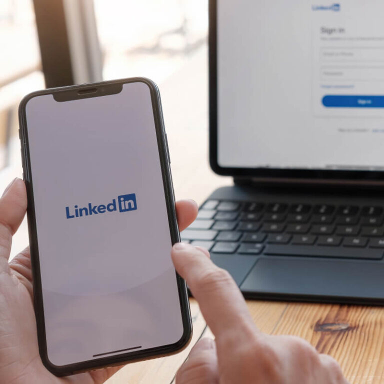 Osoba trzymająca w dłoniach telefon z logo LinkedIn na tle laptopa