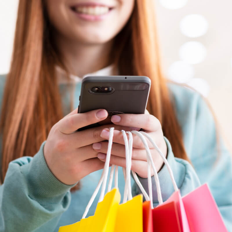 Osoba z rudymi włosami, trzymająca w dłoni telefon i torby na zakupy