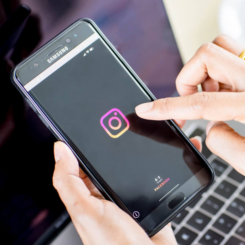 Osoba trzymająca telefon z logo aplikacji Instagram na tle klawiatury laptopa