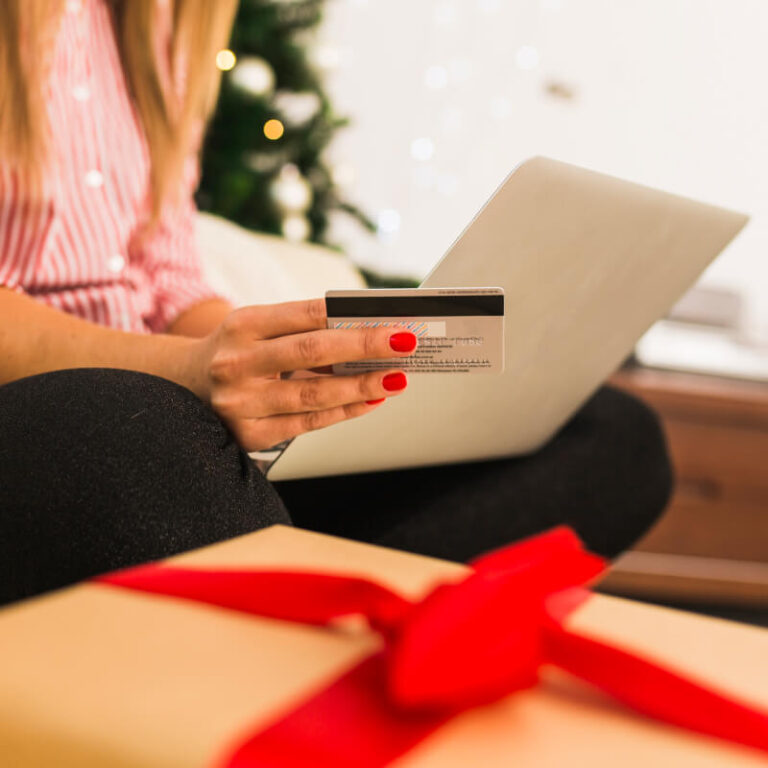 Osoba robiąca świąteczne zakupy online, z kartą płatniczą w dłoni i zapakowanym prezentem na pierwszym planie